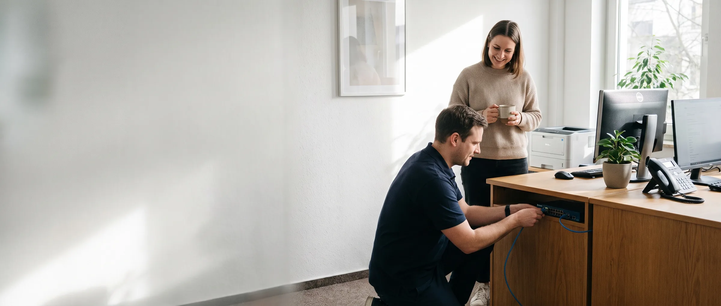 IT-Techniker schließt einen Netzwerk-Switch unter einem Schreibtisch an, eine Mitarbeiterin schaut zu